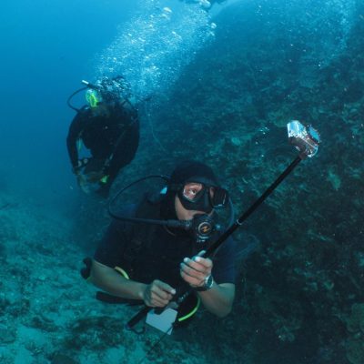 Scuba diver holding a camera on a selfie stick underwater, with another diver in the background near a coral wall.