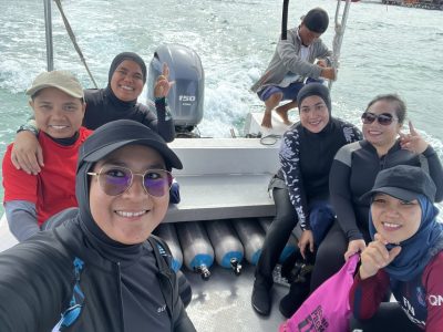 Group of friends on a boat ride to Mabul Island, smiling and wearing wetsuits and casual caps, with the boat motor visible behind them.