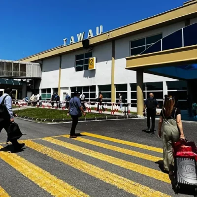 People walking towards the entrance of Tawau Airport on a sunny day, carrying luggage and personal belongings