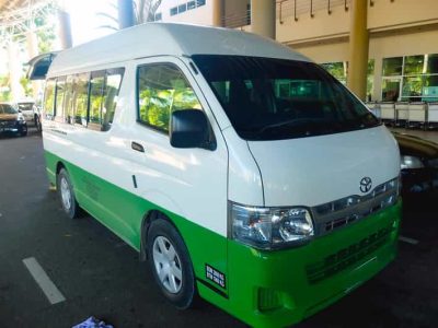 A green and white Toyota van used for airport transfers, parked outside a terminal at Tawau Airport.